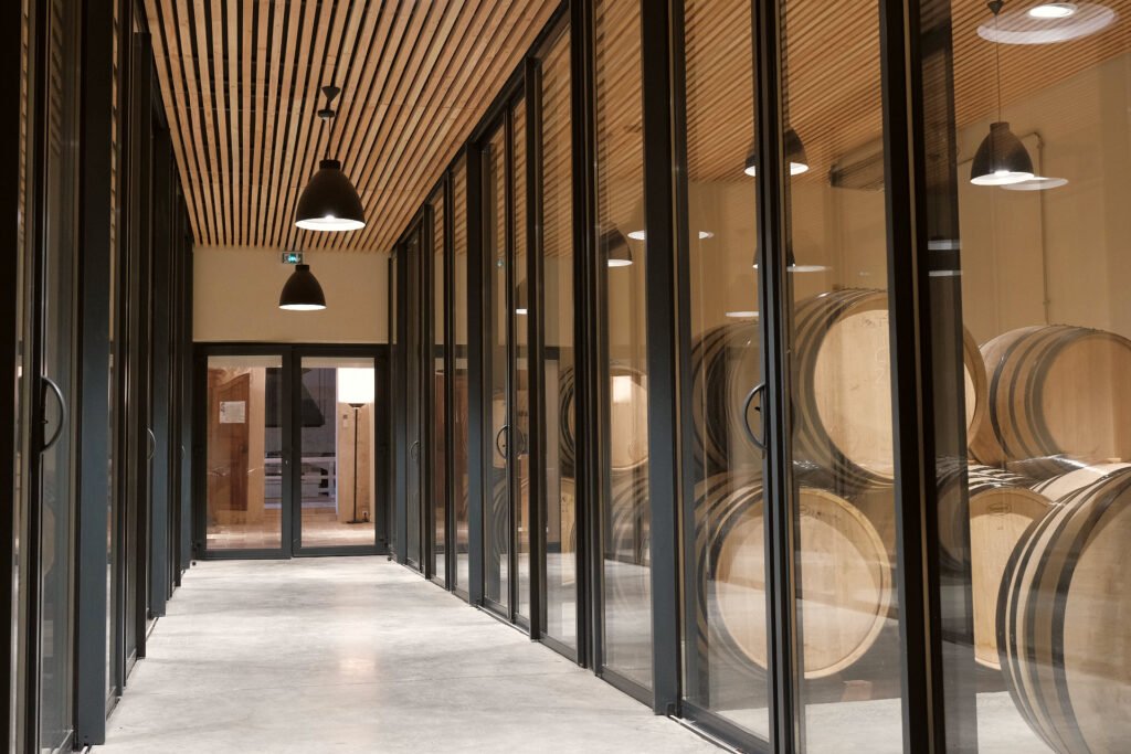 Couloir vitré donnant sur un chai de barriques, au cœur d’un domaine viticole de Bordeaux lors d’une visite œnologique.
