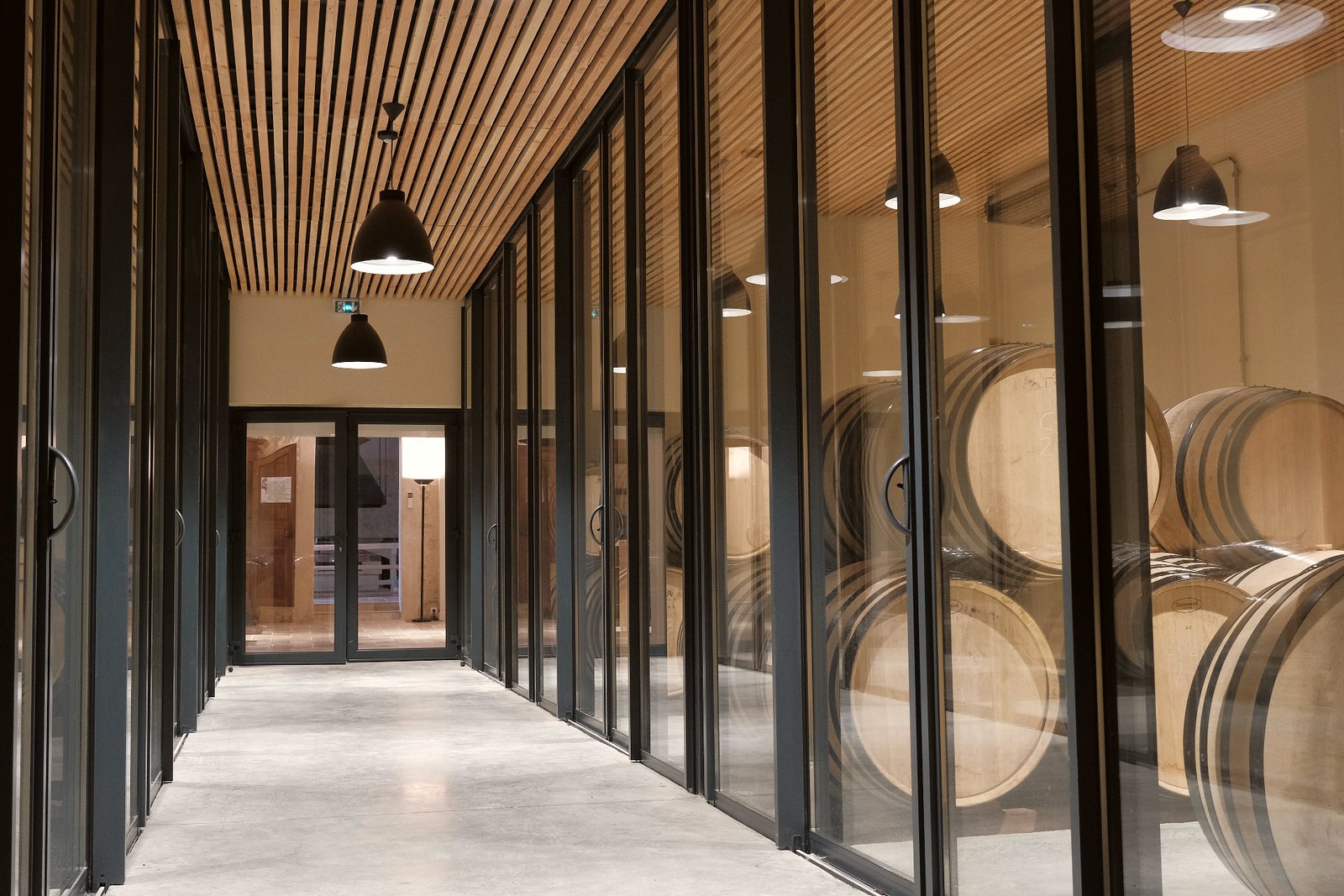 Couloir vitré donnant sur un chai de barriques, au cœur d’un domaine viticole de Bordeaux lors d’une visite œnologique.