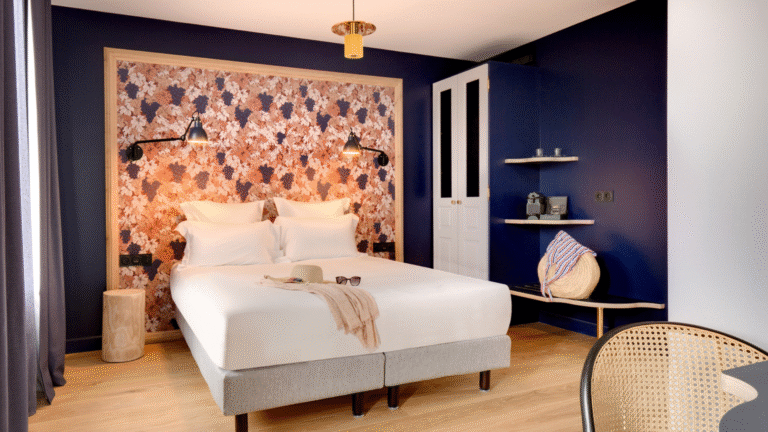 Chambre moderne d’hôtel décorée dans des tons bleu nuit, lit double avec tête de lit à motif floral, éclairage mural et coin café.