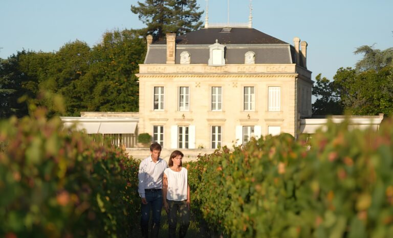 Visite œnotouristique dans les vignes d’un château de Bordeaux, couple marchant devant le domaine au soleil couchant.